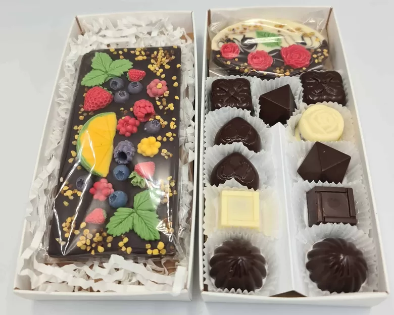 Dāvanu komplekts ar tumšās šokolādes tāfelīti, dekorētu ar ogām un bišu ziedputekšņiem, un dažādām šokolādes konfektēm baltā kastītē.